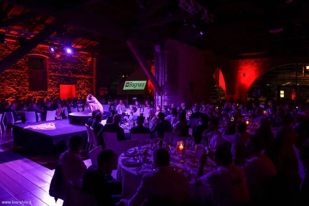 Eine schwach beleuchtete Veranstaltung mit violetter und roter Beleuchtung zeigt Menschen, die an runden Tischen sitzen und eine Duo-Akrobatik-Show auf einer zentralen Bühne verfolgen. Auf einer Leinwand im Hintergrund ist ein Schild mit der Aufschrift "Bagnaca" zu sehen.