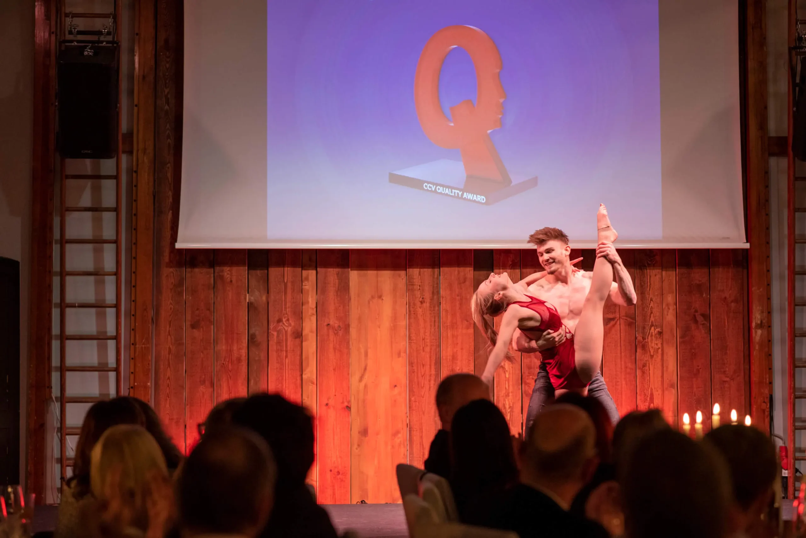 Ein Tänzer und eine Tänzerin führen auf der Bühne vor dem Publikum eine akrobatische Pose auf, um ihre Duo-Akrobatik-Show zu präsentieren. Hinter ihnen ist ein Bildschirm mit einem großen roten Q und der Aufschrift QIX QUALITY AWARD zu sehen.