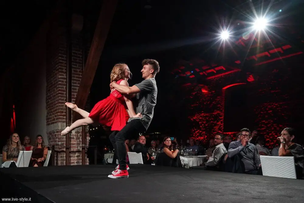 In einer schillernden Duo-Akrobatik-Show tanzen ein Mann und eine Frau energisch zusammen auf der Bühne; sie trägt ein rotes Kleid und wird von ihm hochgehoben, während die Zuschauer von den Tischen im warm beleuchteten Veranstaltungsraum aus aufmerksam zusehen.