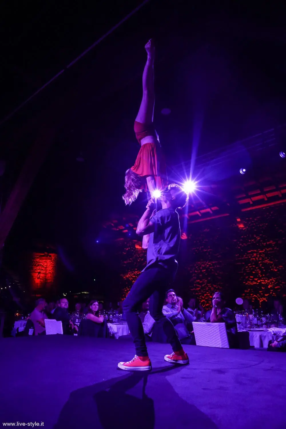 Ein männlicher Akrobat balanciert seine Partnerin während einer Duo-Akrobatik-Show kopfüber auf seiner Schulter, mit dramatischer violetter Beleuchtung und einem an Tischen sitzenden Publikum im Hintergrund.