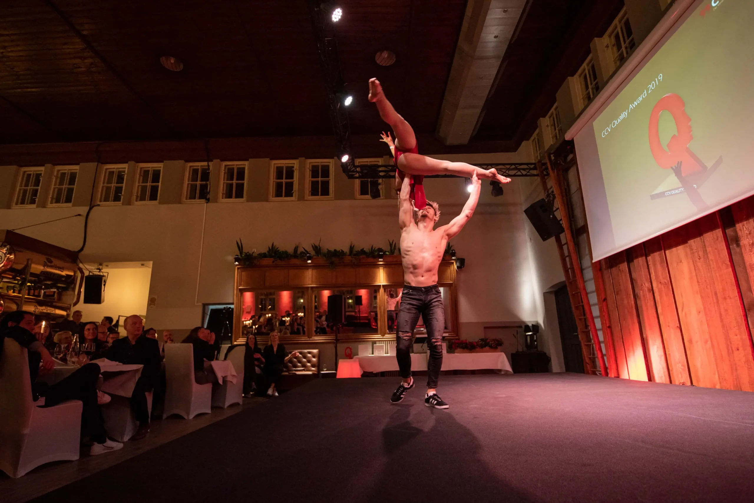 Während einer Duo-Akrobatik-Show hebt ein männlicher Akrobat eine weibliche Person hoch über seinen Kopf und fesselt damit das Publikum, das an Tischen in einem warm beleuchteten Veranstaltungsraum sitzt.