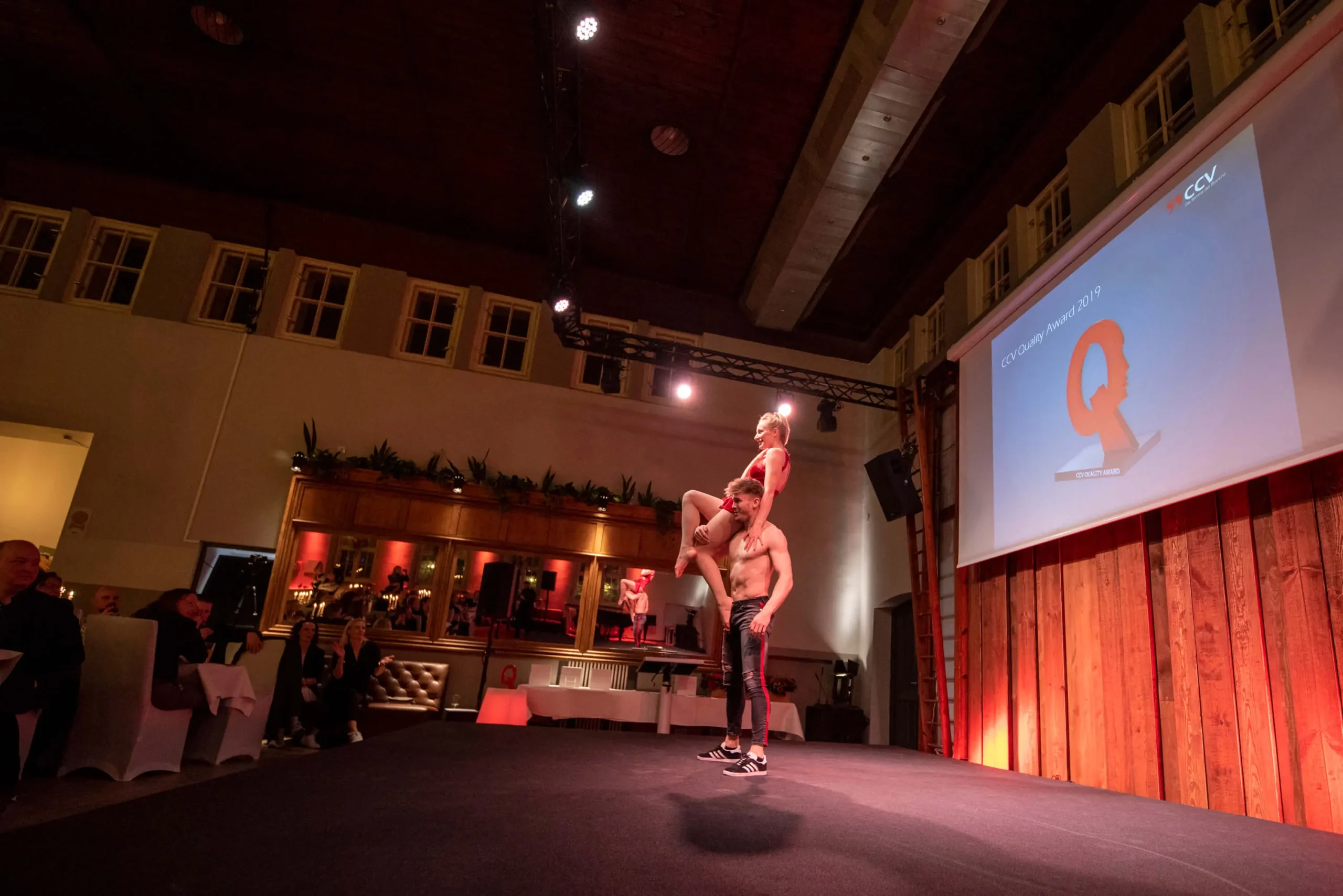 Drei Akrobaten führen eine Duo-Akrobatik-Show auf einer Hallenbühne auf, wobei einer steht und zwei andere balancieren. Das Publikum schaut zu, während im Hintergrund eine Leinwand mit dem großen Buchstaben Q und dem CCV-Logo erscheint.