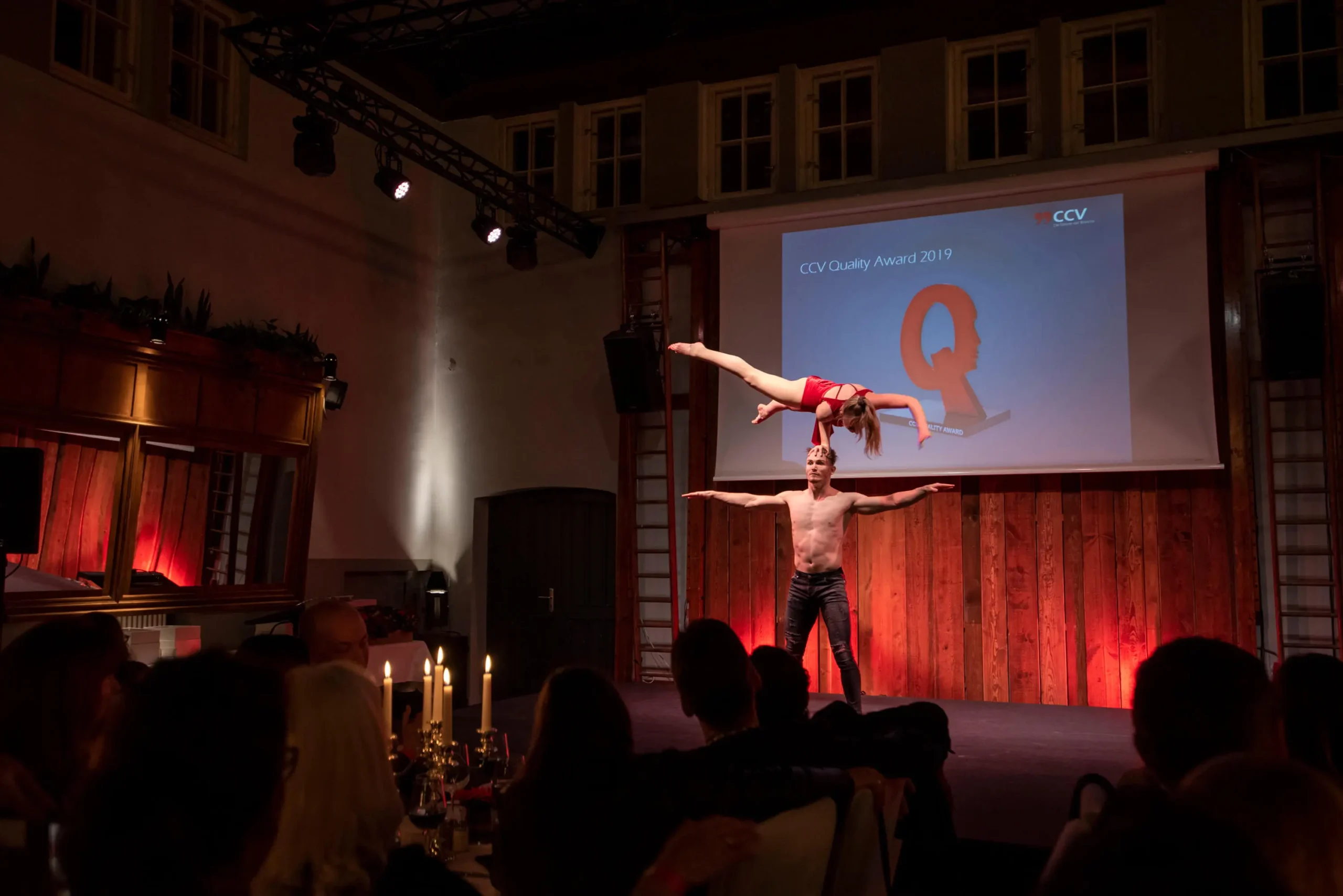 Zwei Akrobaten führen auf der Bühne eine Duo-Akrobatik-Show auf; ein Mann balanciert mit ausgestreckten Armen, während eine Frau anmutig auf einer Hand posiert. Das Publikum schaut zu, während auf einem Bildschirm im Hintergrund der CCV Quality Award 2019 angezeigt wird.