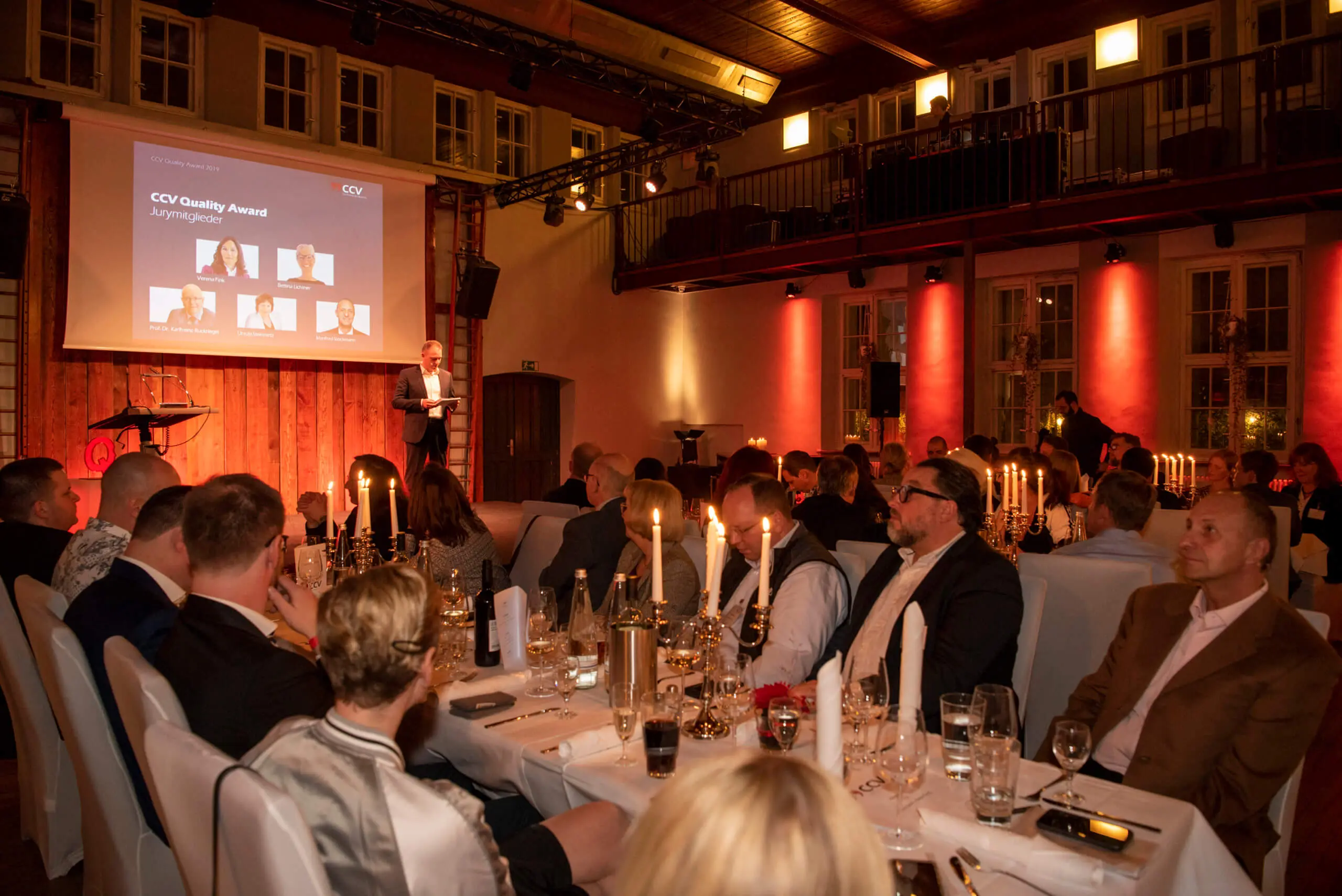 Eine formelle Veranstaltung, bei der die Gäste an Tischen mit Kerzenlicht sitzen und einem Redner auf der Bühne zuhören. Auf einem großen Bildschirm hinter dem Redner werden Informationen über eine Qualitätsauszeichnung angezeigt, während der Raum in warmes Licht getaucht wird - die Gäste warten auf eine beeindruckende Duo Akrobatik Show.