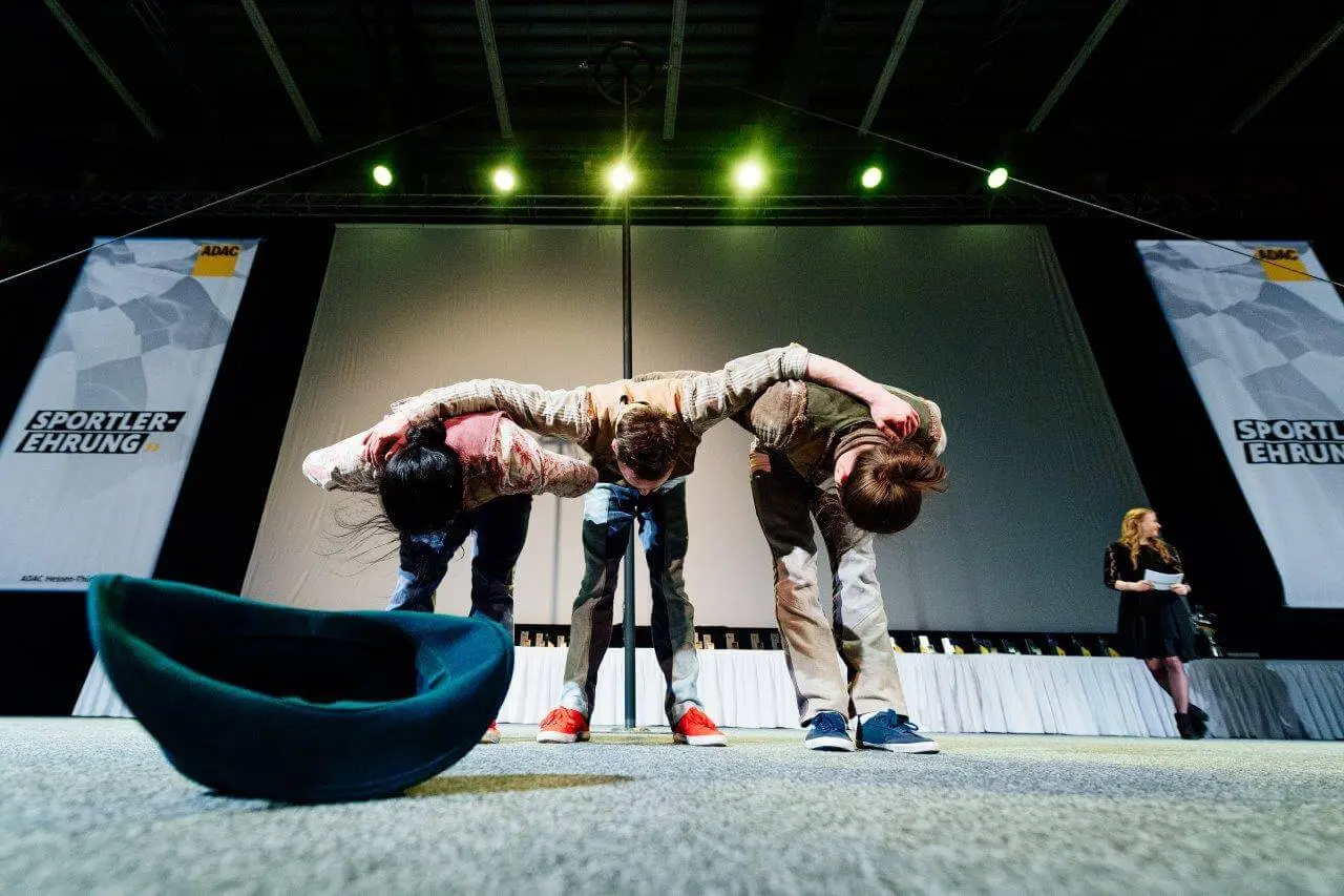 Drei Personen auf der Bühne verbeugen sich unter grellem Licht mit den Armen um die Schultern, während eine Frau an der Seite steht und im Hintergrund zwei Banner mit dem Text Sportler-Ehrung hängen. Vorne auf dem Boden liegt ein Comedy-Artistik-Hut.
