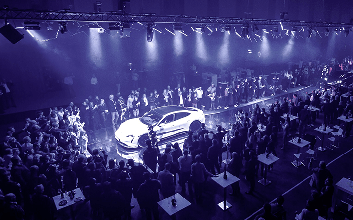 Eine große Menschenmenge versammelt sich um ein hell erleuchtetes Auto, das auf einer Bühne bei einer Indoor-Veranstaltung ausgestellt ist. Scheinwerfer, erhöhte Beleuchtung und eine beeindruckende LED-Show schaffen eine dramatische Atmosphäre. Tische und Teilnehmer umgeben das Auto.