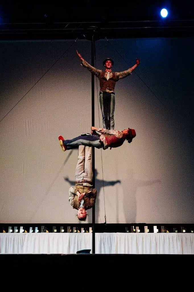 Drei Comedy Artistik-Darsteller in farbenfrohen Kostümen balancieren vertikal auf einer hohen Stange auf der Bühne, einer oben, einer kopfüber in der Mitte und ein weiterer kopfüber unten, alles beleuchtet von einem hellen Scheinwerfer.