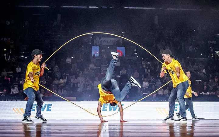 Drei Personen in gelben Hemden führen während einer Halbzeit-Show auf der Bühne eine doppelte holländische Springseilnummer auf; einer tanzt auf den Händen, während die anderen die Seile drehen, während das Publikum im Hintergrund zusieht.