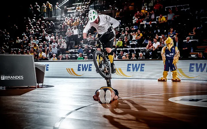 Während einer aufregenden Halbzeit-Show vollführt ein Radfahrer einen Stunt, indem er mit dem Fahrrad über eine Person springt, die mit dem Gesicht nach unten auf einem Basketballfeld liegt, während die Menge zusieht und ein kostümiertes Maskottchen in der Nähe steht.