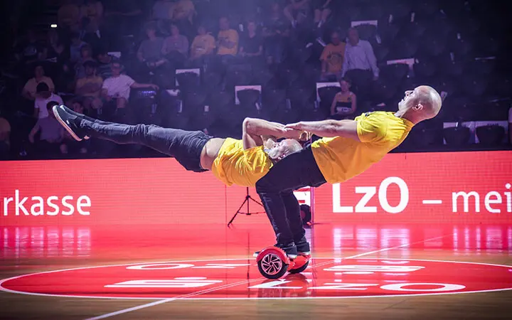 Zwei Männer in gelben Hemden vollführen während einer Halbzeit-Show in einer hell erleuchteten Halle einen waghalsigen Balanceakt auf einem Hoverboard, wobei sich ein Mann zurücklehnt, während er den anderen waagerecht hält. Im Hintergrund schaut ein Publikum zu.