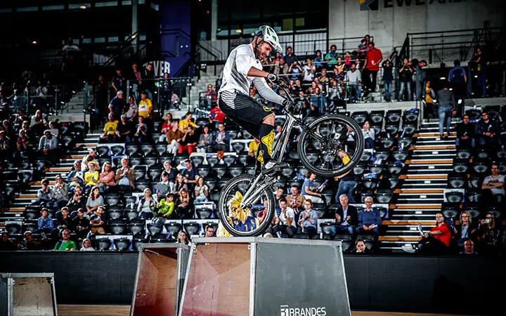 Ein Radfahrer, der einen Helm trägt, führt ein Kunststück auf einem Fahrrad aus und balanciert dabei auf dem Hinterrad auf einer Box in einer Arena während der Halbzeit-Show, während die Zuschauer von der Tribüne im Hintergrund zusehen.