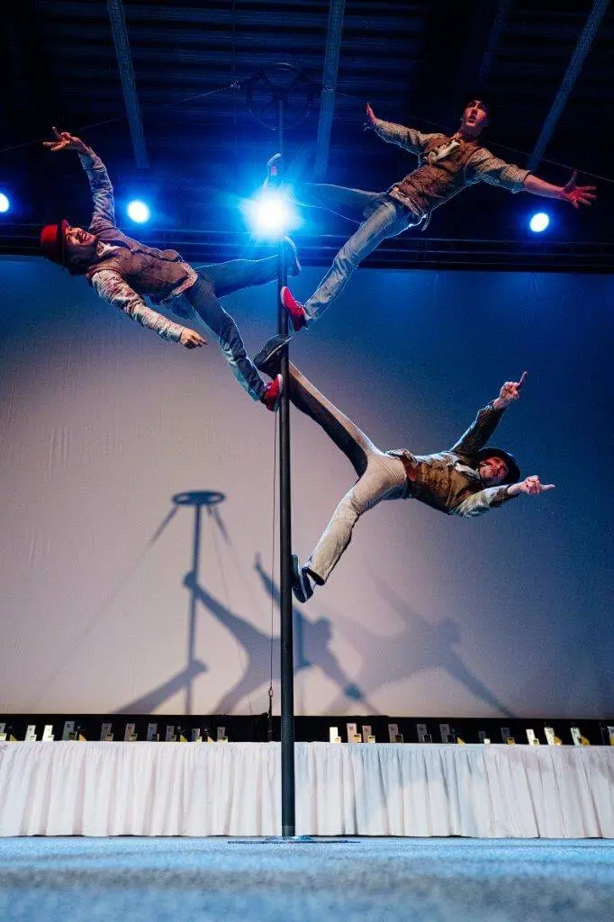 Drei Darsteller in aufeinander abgestimmten Outfits balancieren horizontal auf einer vertikalen Stange, wobei jeder einen Arm und ein Bein ausstreckt und Comedy-Artistik mit akrobatischem Geschick verbindet, während die dramatische Bühnenbeleuchtung auffällige Schatten auf die Kulisse hinter ihnen wirft.