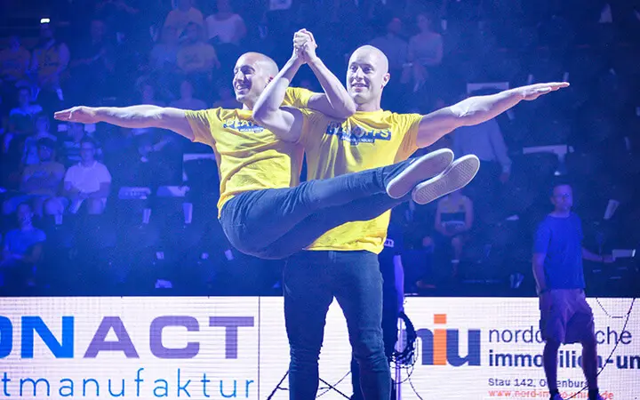Zwei Männer in passenden gelben Hemden führen während einer Halbzeit-Show eine akrobatische Hebefigur auf der Bühne aus, wobei der eine den anderen mit ausgestreckten Armen waagerecht hält. Im dunklen Hintergrund sind Leuchtreklamen und ein sichtbares Publikum zu sehen.