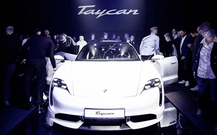 Ein weißer Porsche Taycan wird bei einer Veranstaltung im Scheinwerferlicht ausgestellt, umgeben von Menschen, die Fotos machen und das Auto bewundern. Eine beeindruckende LED-Show beleuchtet das große Taycan-Logo auf einem Bildschirm im Hintergrund.