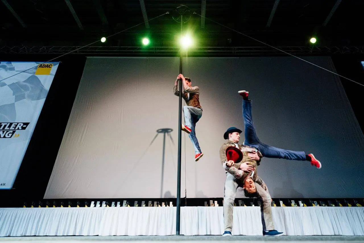 Drei Comedy-Artistik-Darsteller stehen auf der Bühne; einer klettert auf eine senkrechte Stange, während die beiden anderen, die Hüte tragen, eine akrobatische Bewegung ausführen, wobei einer den anderen kopfüber an der Taille hält. Hinter ihnen ist eine große weiße Leinwand zu sehen.