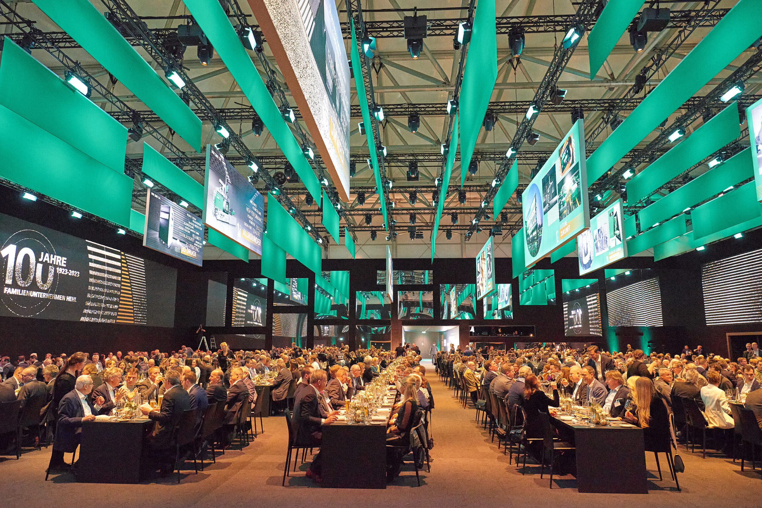 Virtuelle Events Stuttgart: In einem geräumigen, modernen Veranstaltungssaal mit heller türkisfarbener Beleuchtung und von der Decke hängenden Digitalanzeigen sitzt eine große Menge formell gekleideter Menschen an langen Tischen.