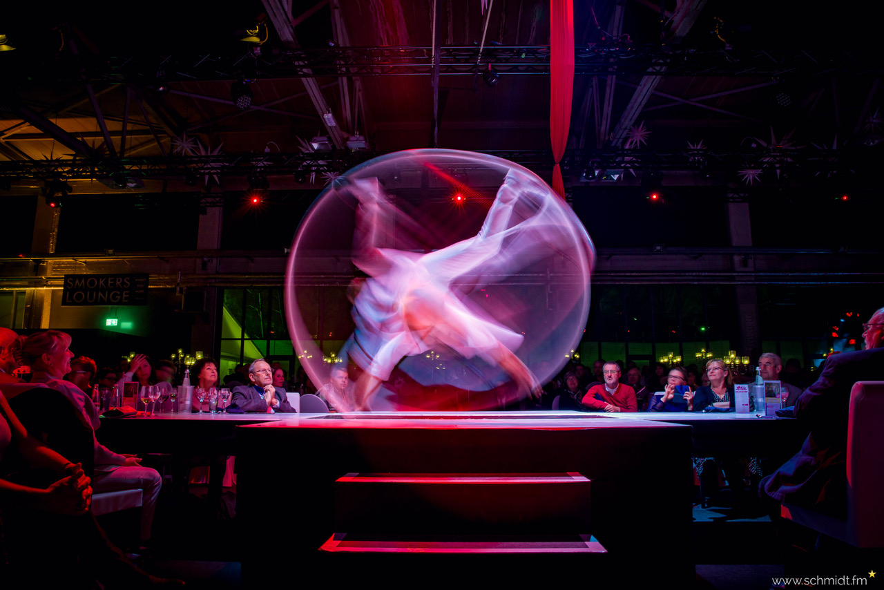 Ein Zirkusartist dreht sich schnell in einer großen durchsichtigen Kugel auf der Bühne, wodurch ein verschwommener Bewegungseffekt entsteht, während das Publikum an Tischen sitzt und unter farbenfroher, dramatischer Beleuchtung zusieht.