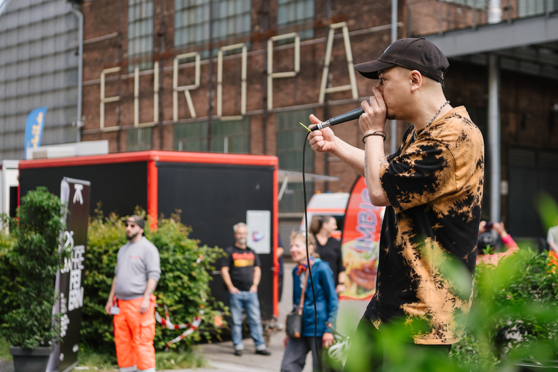 Ein Mann in einem schwarz-orangefarbenen Krawattenhemd spricht im Freien mit einem Mikrofon, während eine kleine Gruppe von Menschen in der Nähe eines Gebäudes mit der Aufschrift EUROPA zuhört. Im Hintergrund sind Büsche und ein roter Wohnwagen zu sehen.