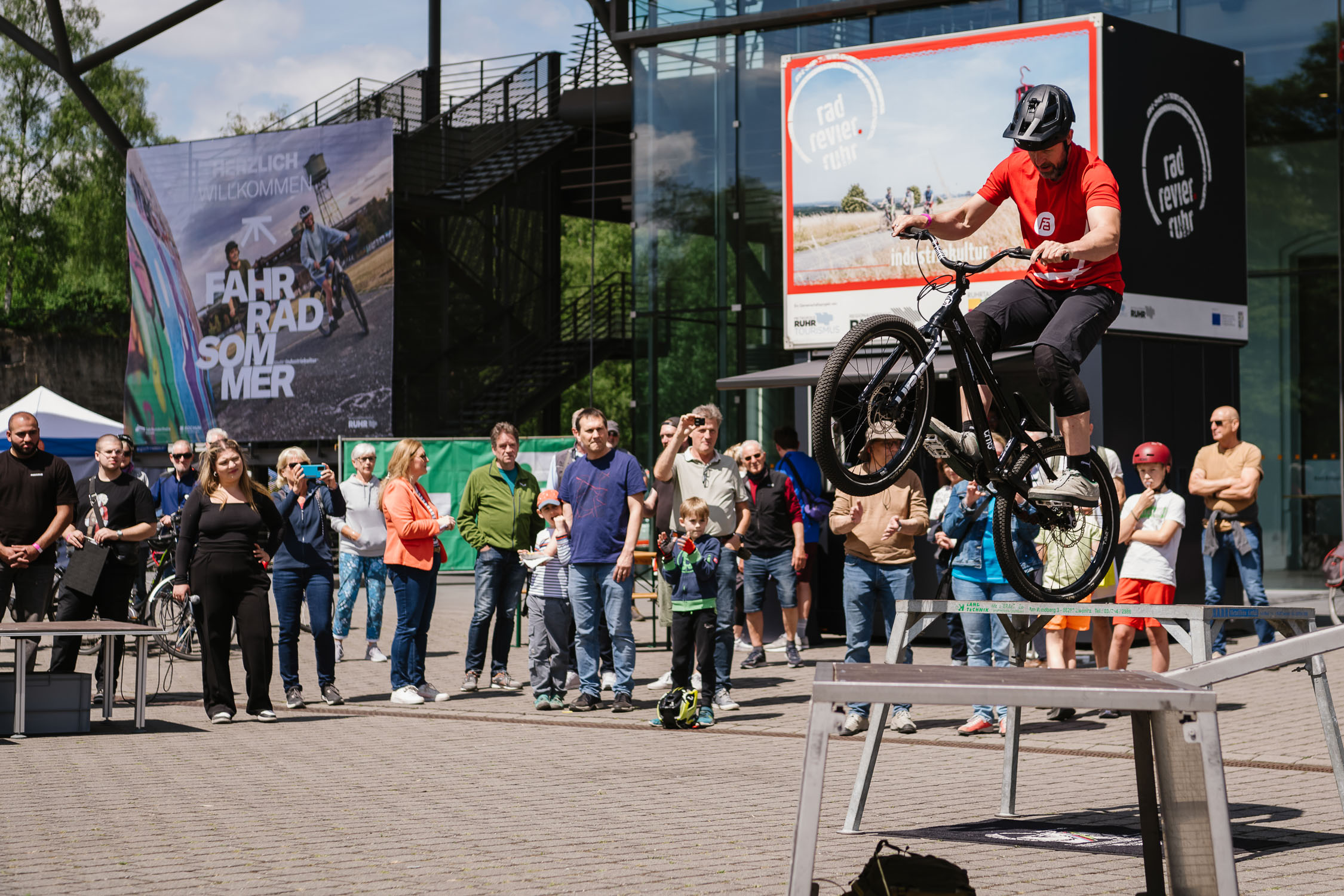 Ein Radfahrer vollführt in der Luft einen Stunt auf einem Fahrrad über einer Bank, während eine Gruppe von Erwachsenen und Kindern an einem sonnigen Tag im Freien in der Nähe eines modernen Gebäudes zusieht.