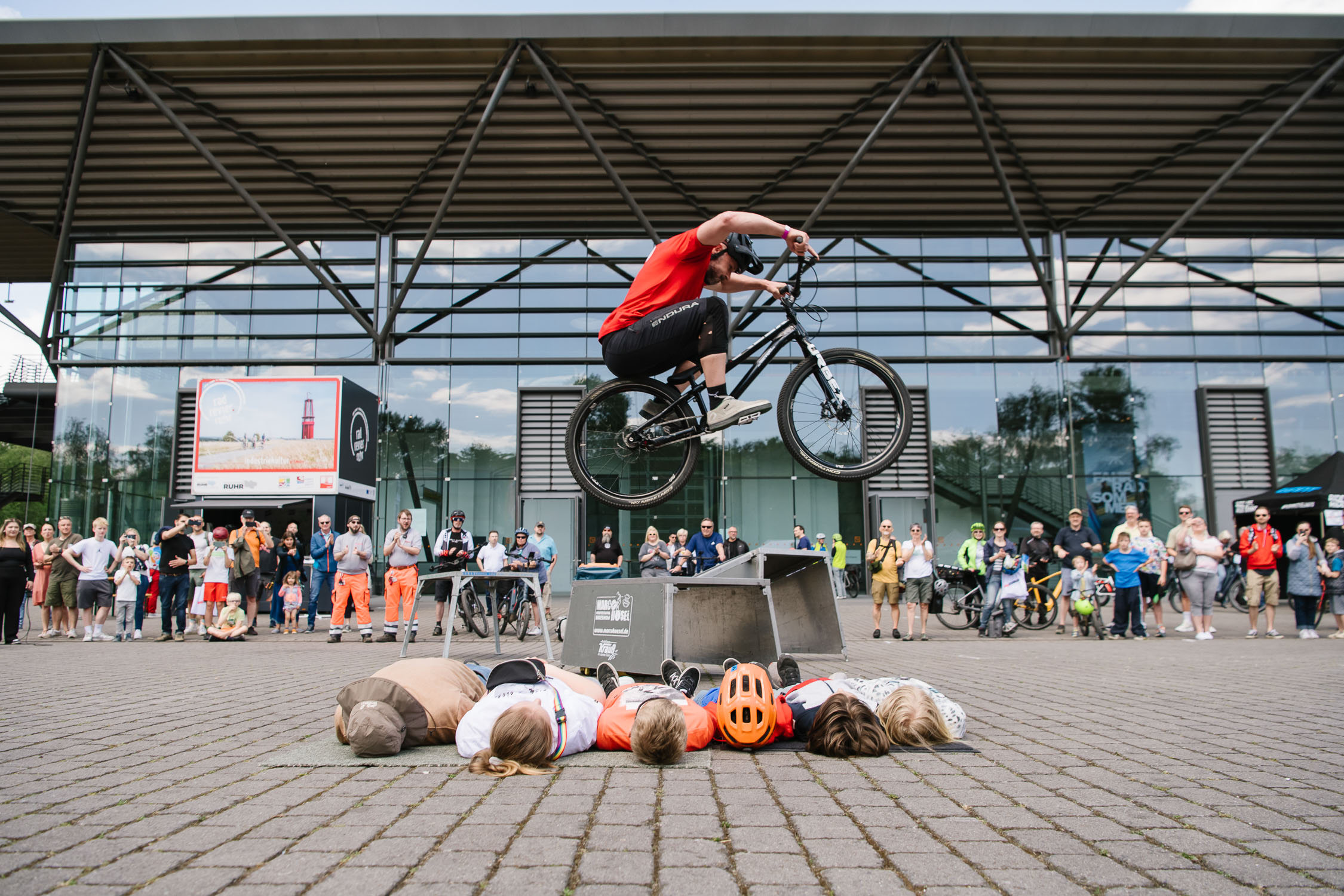 Ein Radfahrer springt mit einem BMX-Rad in der Luft über vier Personen, die nebeneinander auf dem Boden liegen, während eine Menge von Zuschauern vor einem modernen Glasgebäude zusieht.