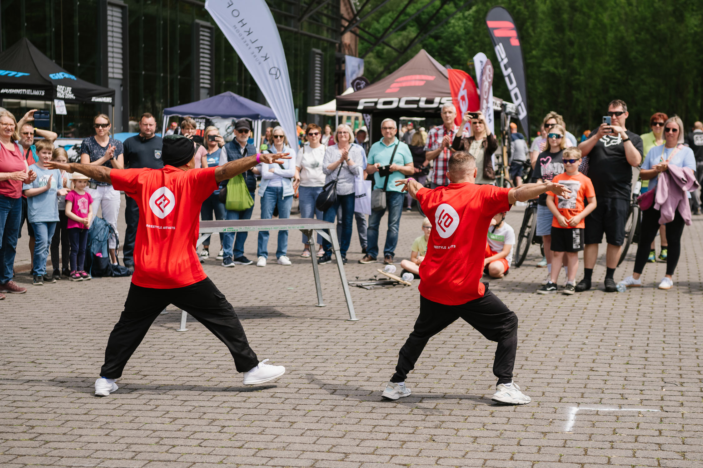 Zwei Darsteller in passenden roten Hemden und schwarzen Hosen tanzen energisch vor einer Menschenmenge im Freien, während Schaulustige in der Nähe von Veranstaltungszelten und Bannern die Aufführung beobachten und aufnehmen.