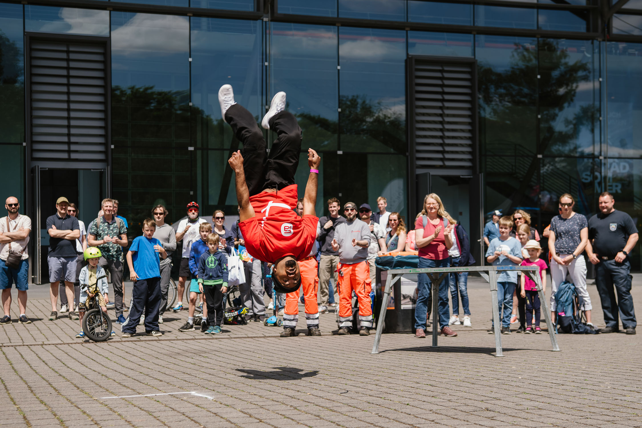 Ein Mann in einem roten Hemd vollführt vor einer Menschenmenge im Freien einen Rückwärtssalto. Menschen aller Altersgruppen schauen zu, einige lächeln, als der Mann sich in der Luft über dem Bürgersteig befindet, mit einem Tisch in der Nähe. Im Hintergrund ist ein modernes Glasgebäude zu sehen.