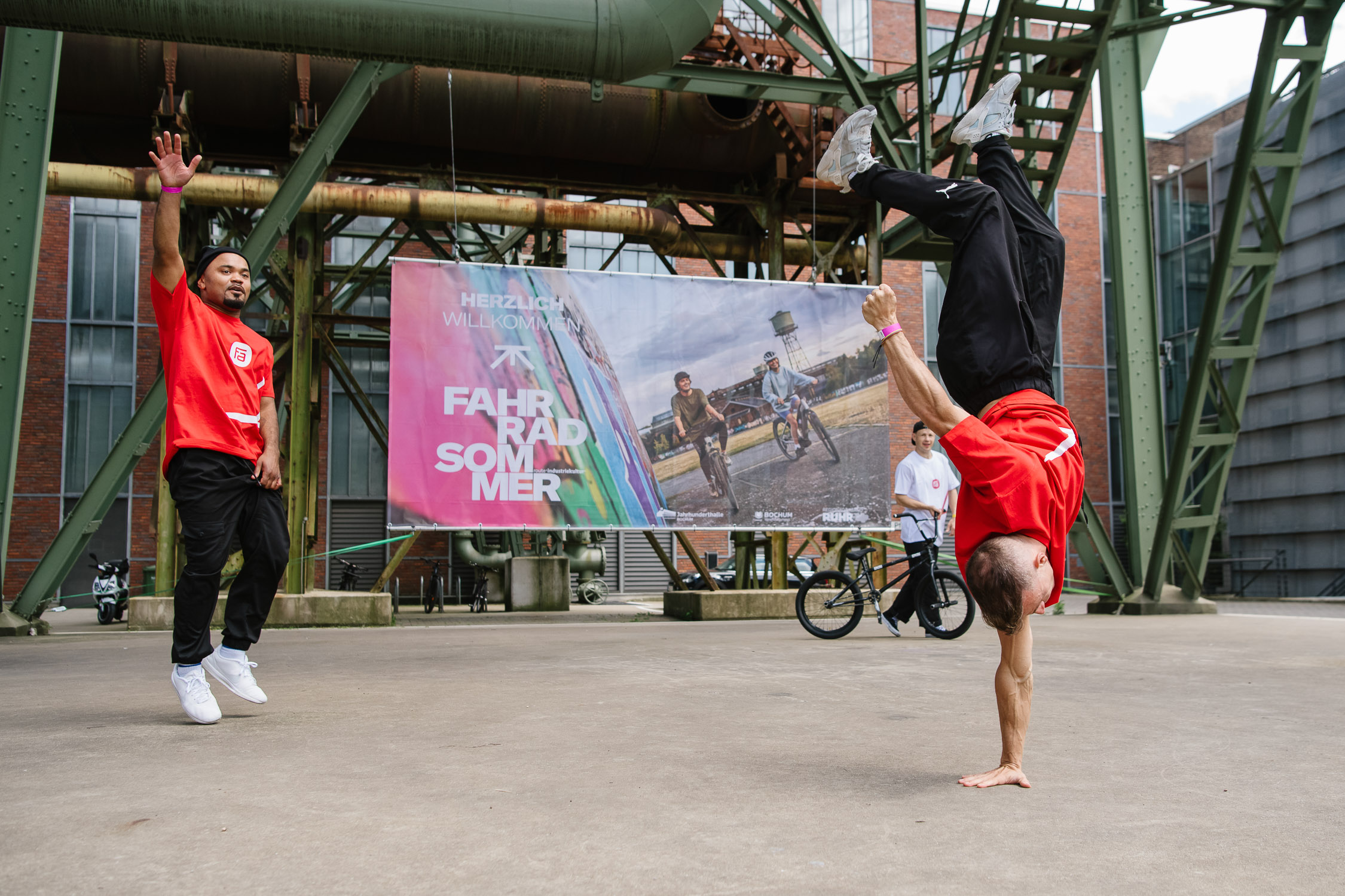 Urban Street Show, Zwei Männer in roten Hemden tanzen Breakdance im Freien unter einer Metallkonstruktion; einer macht einen Handstand, während der andere zusieht. Im Hintergrund sind ein Fahrrad und ein buntes Veranstaltungsbanner zu sehen.