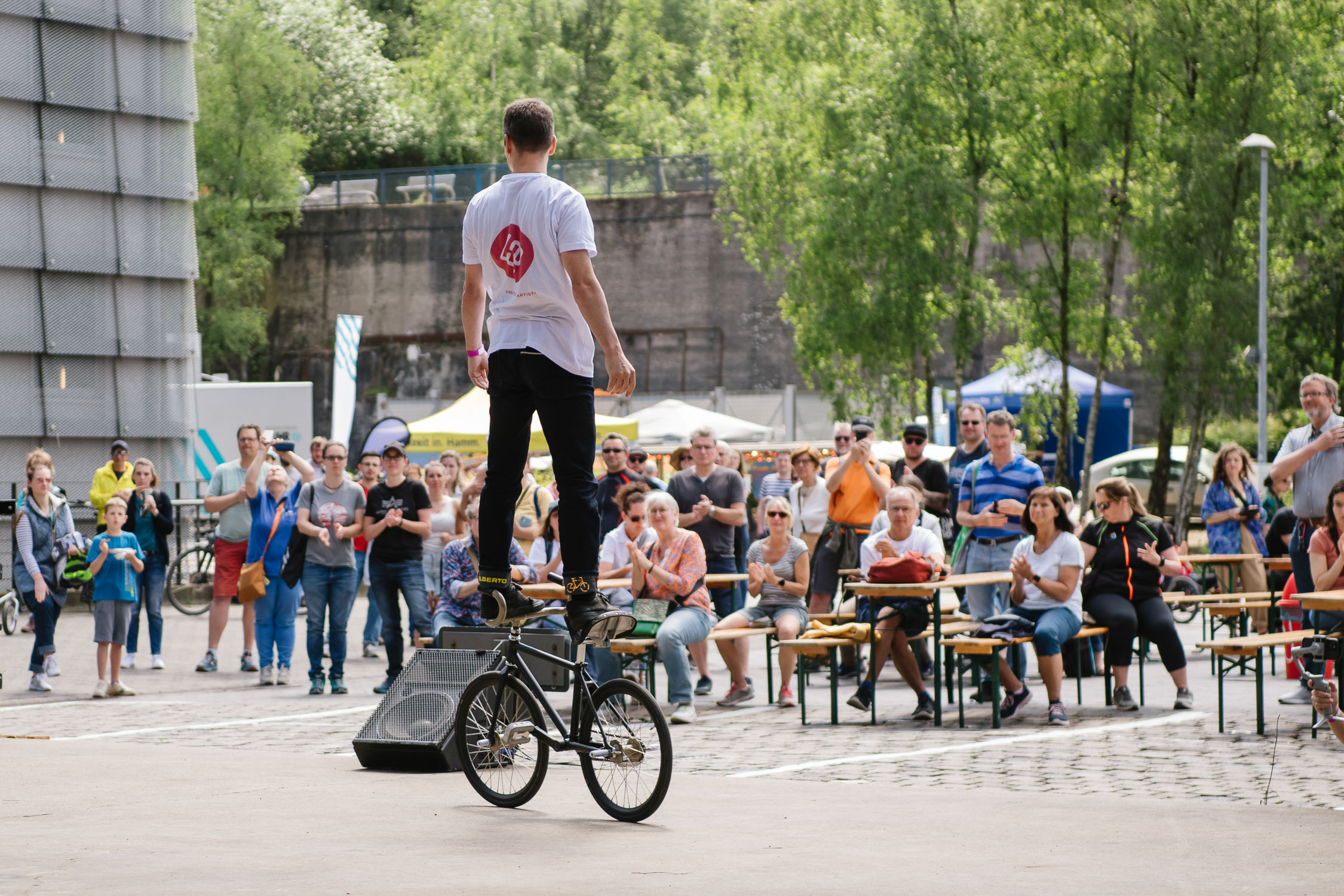 Ein Mann balanciert auf einem Fahrradsattel stehend, während er bei einer Freiluftveranstaltung vor einem Publikum auftritt. Die Zuschauer schauen aufmerksam zu, einige sitzen auf Bänken, im Hintergrund sind Bäume und Zelte zu sehen.