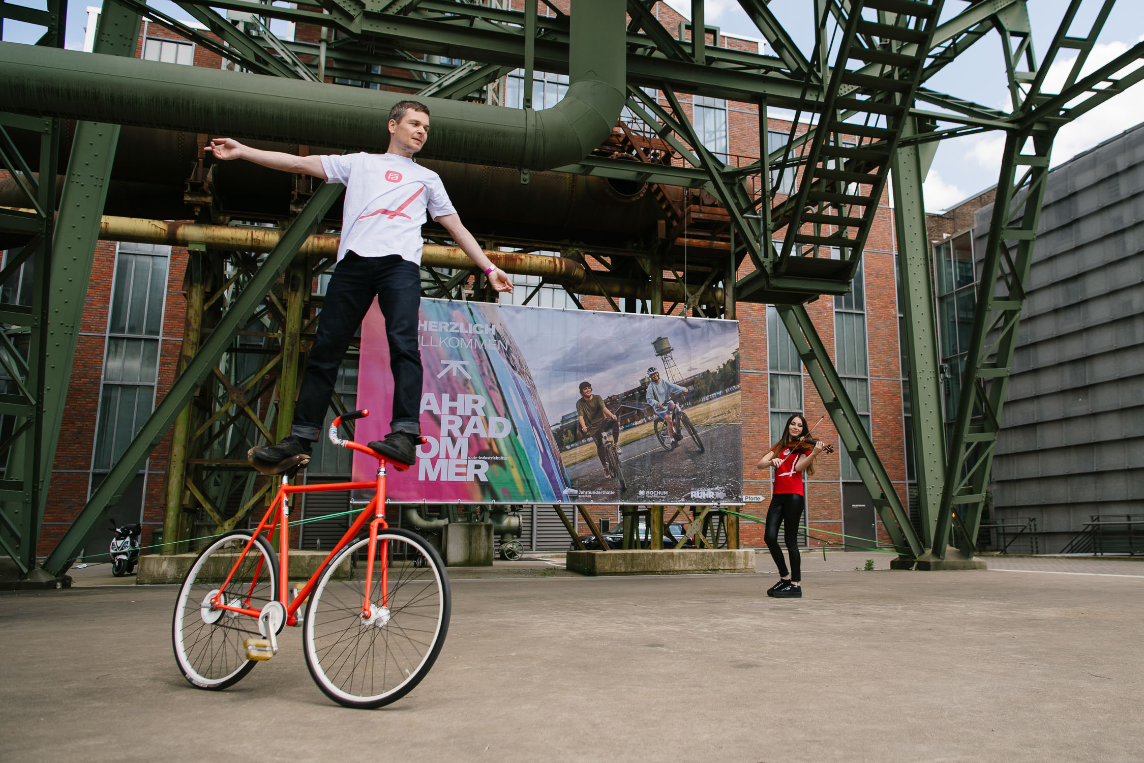 Ein Mann balanciert mit ausgestreckten Armen auf dem Lenker eines roten Fahrrads, während eine Frau in Rot in einer industriellen Umgebung mit Rohren und einem großen Plakat im Hintergrund steht.