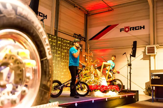 Ein Mann mit einem Mikrofon steht neben zwei BMX-Radfahrern auf der Bühne einer Weihnachtsfeier, mit einem geschmückten Weihnachtsbaum und festlicher Beleuchtung im Hintergrund. Im Vordergrund ist ein großer Reifen zu sehen, der auf die Indoor-Radsportveranstaltung hinweist.