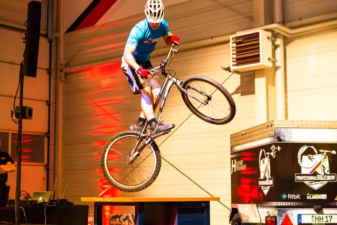 Ein Radfahrer mit Helm und Sportkleidung vollführt einen Sprung auf einem Mountainbike in der Halle, einen Fuß vom Pedal, in der Nähe einer Plattform und eines Markenanhängers - alles unter heller Beleuchtung bei einer festlichen Weihnachtsfeier.