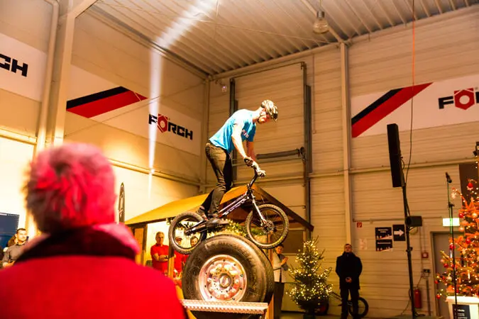Ein Radfahrer in blauem Hemd und Helm vollführt während einer Weihnachtsfeier in einer Halle einen Fahrradstunt auf großen Reifen, während ein Zuschauer in einem roten Mantel zwischen Weihnachtsbäumen und festlichen Lichtern zuschaut.
