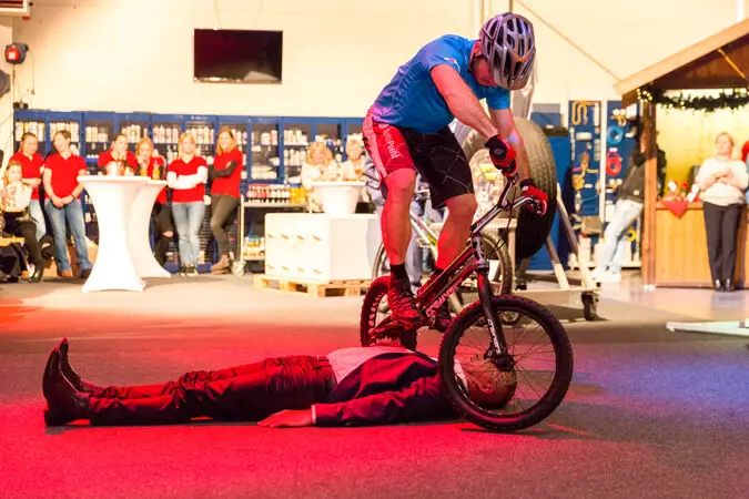 Bei einer Weihnachtsfeier vollführt ein Radfahrer mit Helm und blauem Hemd ein Kunststück, indem er sein Fahrrad auf der Brust eines am Boden liegenden Mannes im Anzug balanciert, während im Hintergrund eine Menschenmenge zusieht.