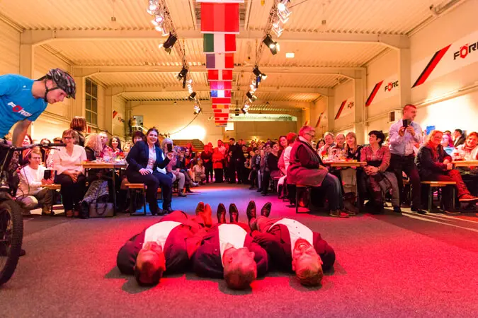 Eine Menschenmenge beobachtet, wie sich ein Radfahrer darauf vorbereitet, über vier Männer in Anzügen zu springen, die bei einer lebhaften Weihnachtsfeier in einer hell erleuchteten und mit hängenden Fahnen geschmückten Halle nebeneinander auf dem Boden liegen.