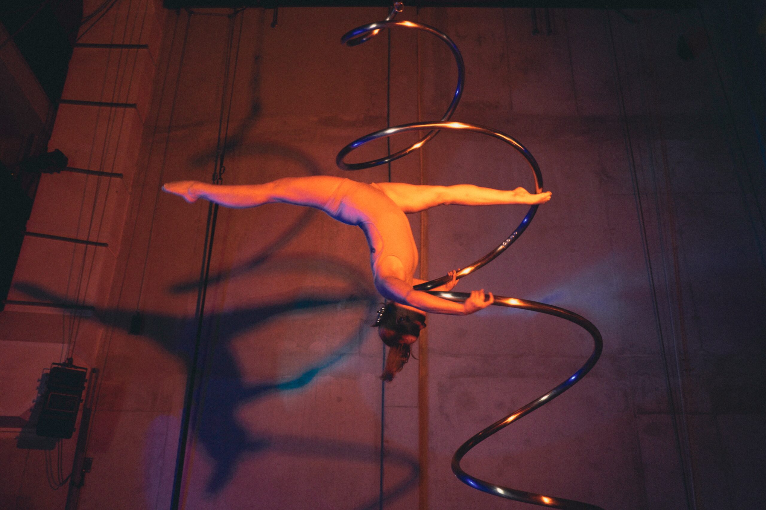 Ein Luftakrobat vollführt einen Spagat, während er an einem großen, spiralförmigen Metallgerät hängt, das auf einer Bühne dramatisch beleuchtet wird.