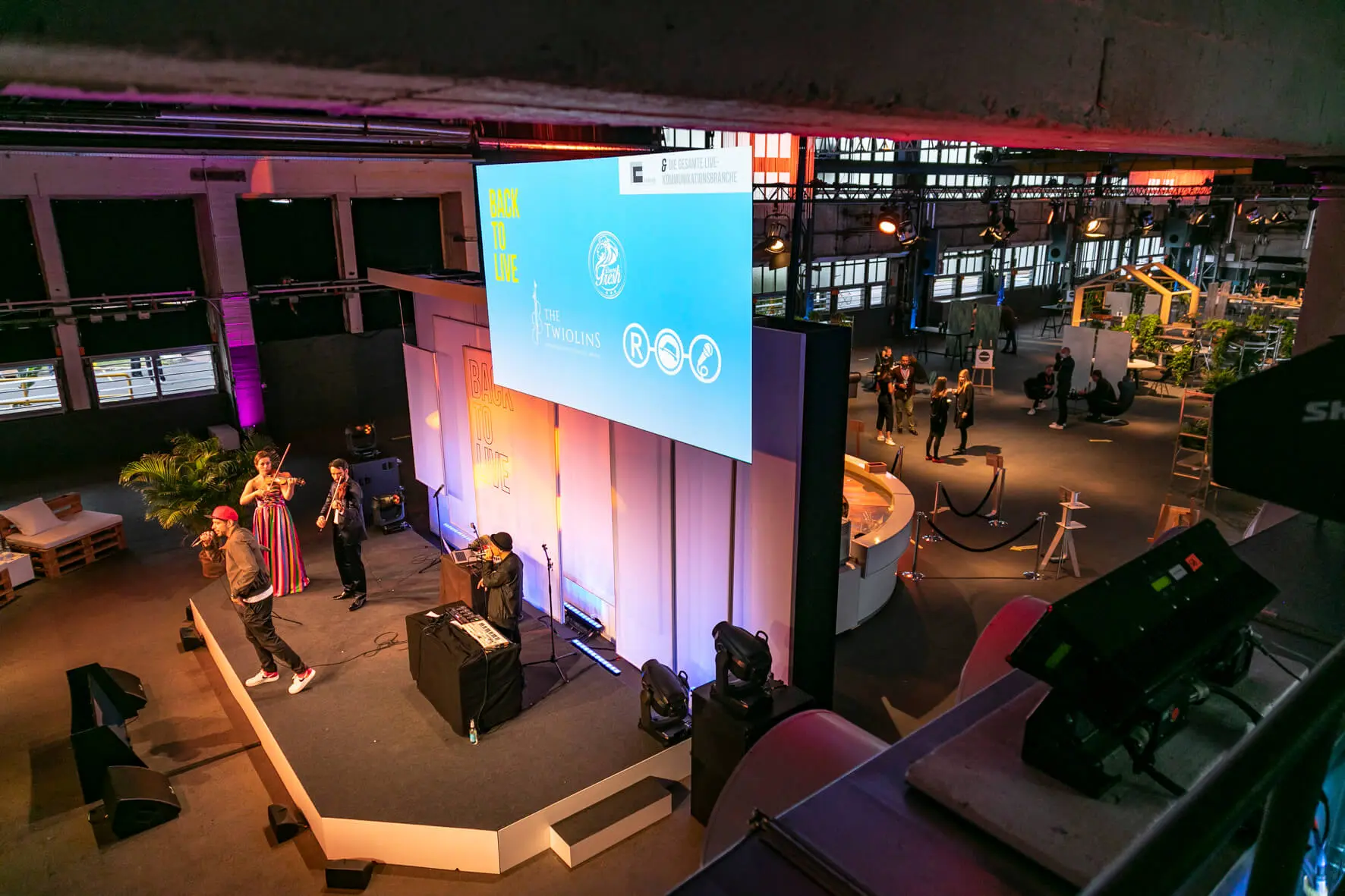 Auf der Back2Live-Veranstaltung steht eine Gruppe von Menschen auf einer Bühne mit Mikrofonen und Ausrüstung in einem großen, industriell anmutenden Raum mit einer großen Leinwand und verstreuten Sitzgelegenheiten und Displays im Hintergrund.