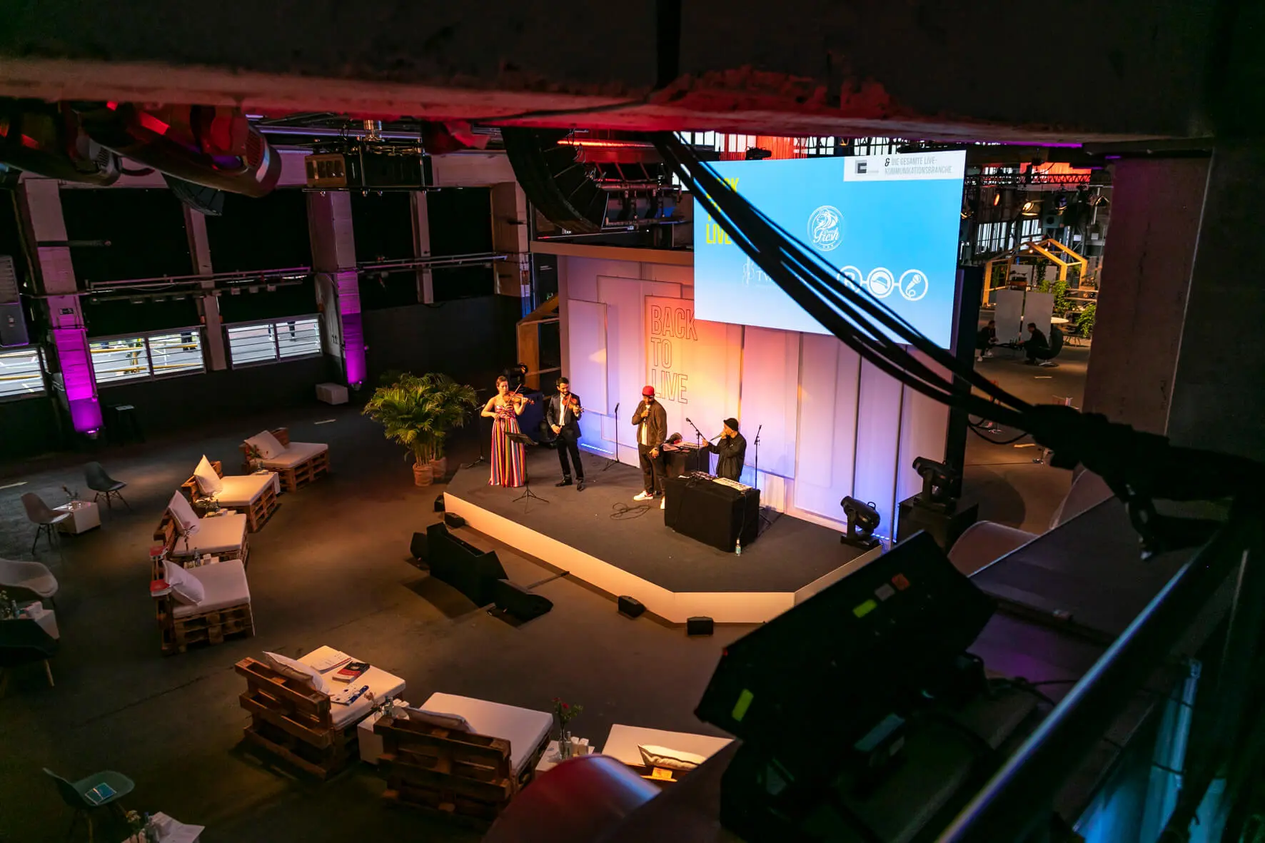 Eine Gruppe von Menschen steht auf einer Bühne mit Mikrofonen im Back2Live, einem modernen Raum im Industriestil, in dem bunte Beleuchtung und Sofas im Hintergrund einen lebendigen Publikumsbereich schaffen.