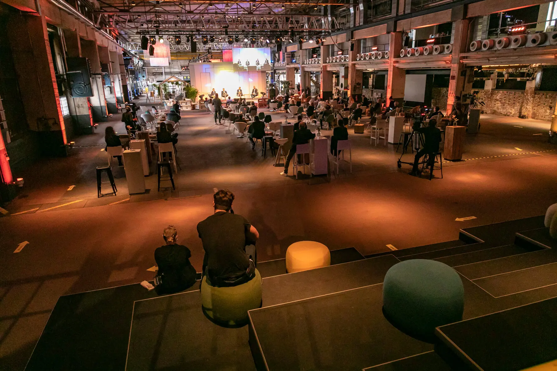 Im Back2Live, einer geräumigen Veranstaltungshalle im Industriestil, sitzen die Gäste an Tischen und auf Hockern und schauen auf eine hell erleuchtete Bühne im vorderen Bereich. Die Atmosphäre ist zwanglos, mit verstreuten Sitzgelegenheiten und warmer Beleuchtung.