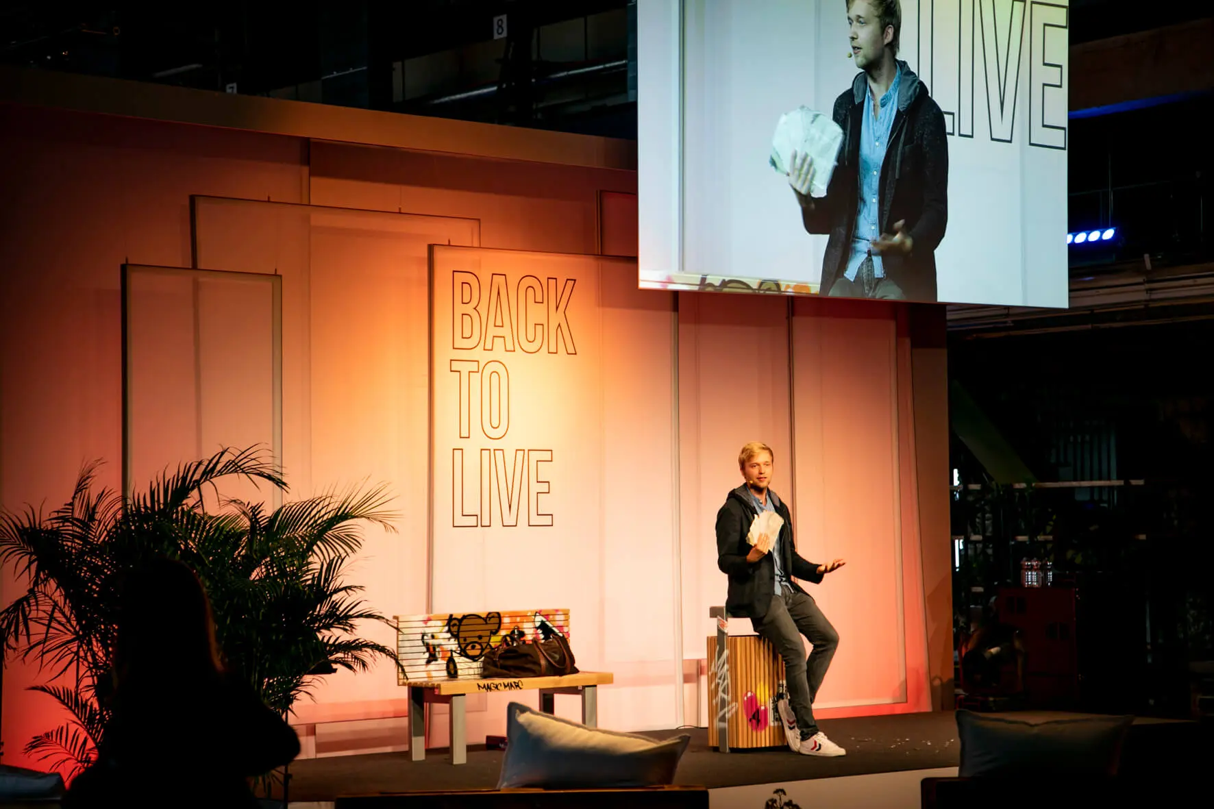 Ein Redner präsentiert auf der Bühne einer Veranstaltung, während Back2Live hinter ihm angezeigt wird. Er hält Papiere in der Hand und sein Bild wird auf eine große Leinwand projiziert. Die moderne, gut beleuchtete Umgebung mit Pflanzen und Sitzgelegenheiten spiegelt die lebendige Atmosphäre von Back2Live wider.