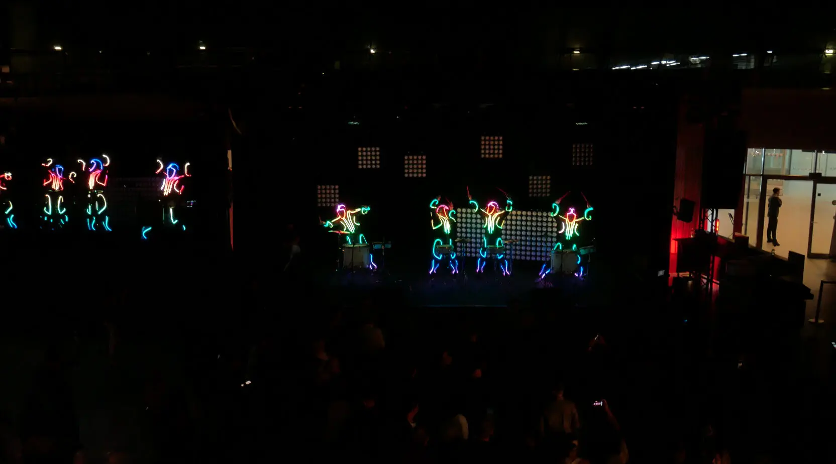 Schwarz gekleidete Performer, die ihre Körper und Hüte mit bunten Neonlichtern schmücken, tanzen auf einer dunklen Bühne und kreieren leuchtende Formen und Muster, die an eine LED-Trommelshow erinnern.