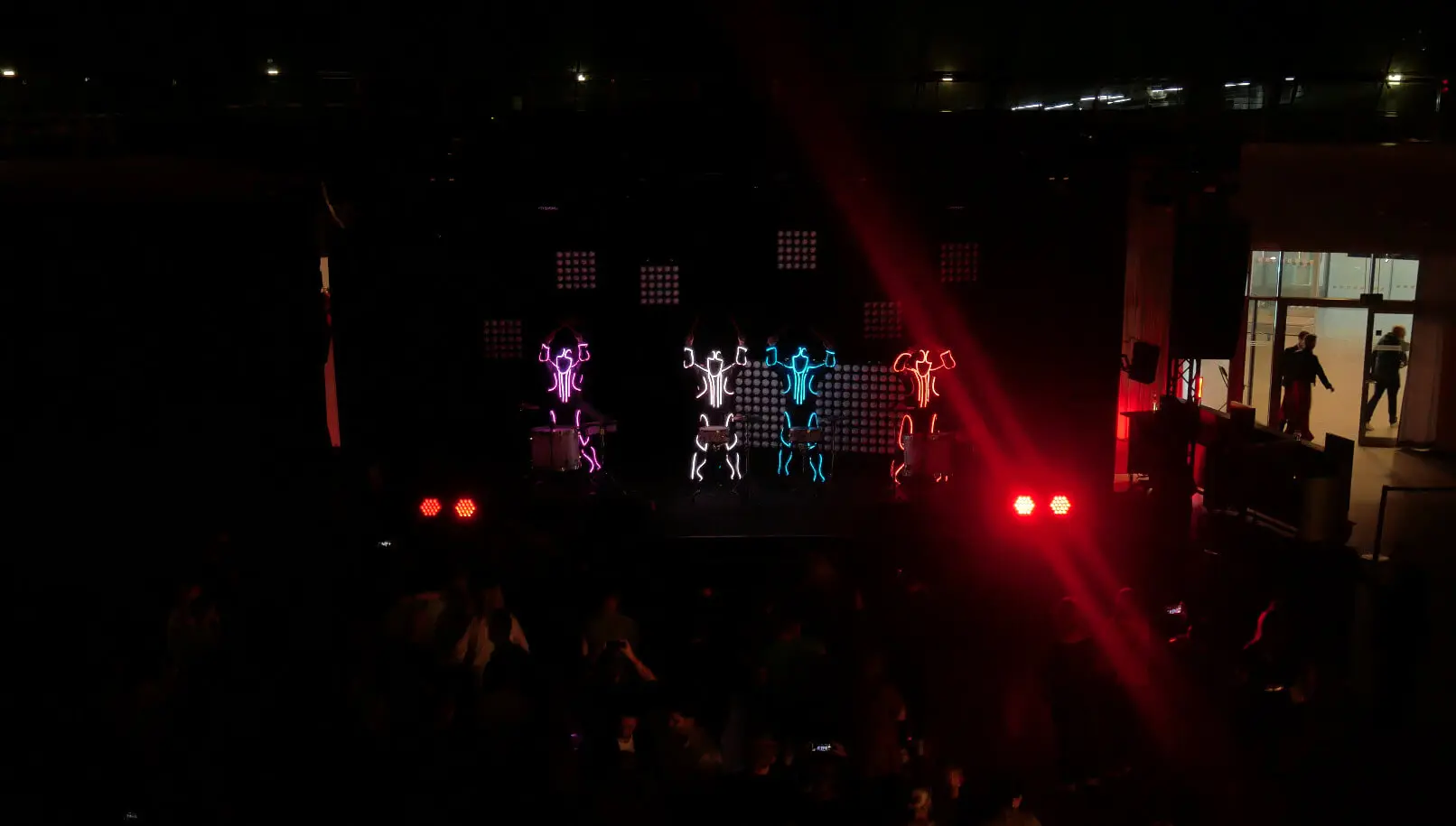 Eine dunkle Bühne mit Darstellern, die in leuchtende Neonlichter gehüllt sind und LED Drum Show Strichmännchen in Pink, Blau und Orange bilden. Rote Scheinwerfer leuchten vorne, und im unteren Vordergrund ist ein schwaches Publikum zu sehen.