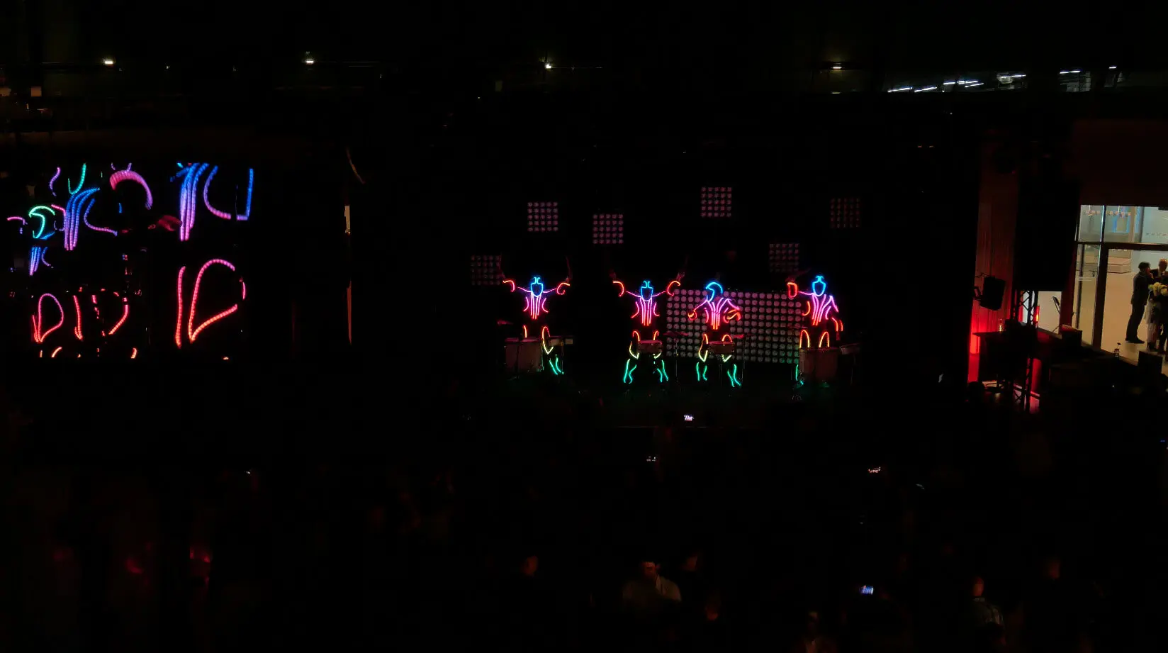 Die Darsteller auf der dunklen Bühne tragen leuchtende, bunte LED-Anzüge in Form stilisierter menschlicher Figuren, die vor einem schwarzen Hintergrund leuchtende LED-Trommel-Show-Muster erzeugen. Auf der rechten Seite sind ein Publikum und ein hell erleuchteter Eingang zu sehen.