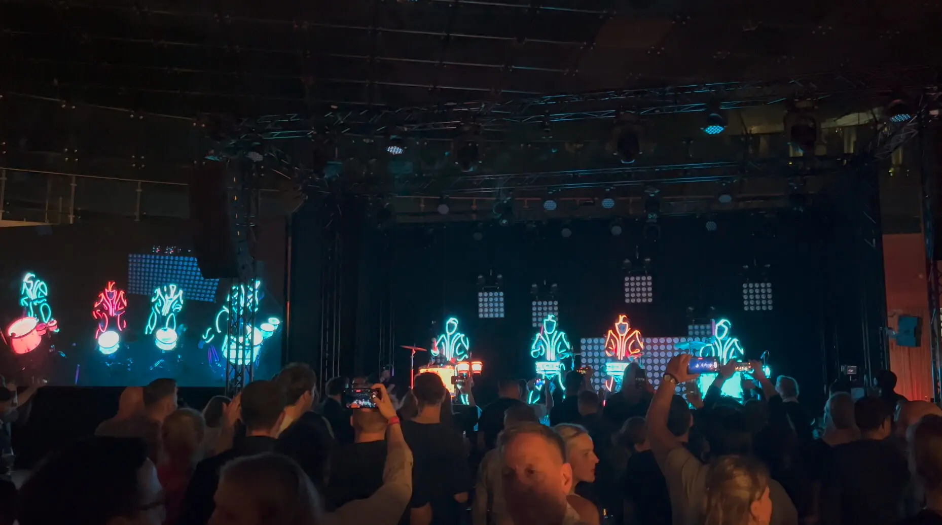 Eine Menschenmenge verfolgt ein Live-Konzert in einer dunklen Halle. Auf der Bühne stehen Musiker in leuchtenden Neonkostümen, die von einer elektrisierenden LED-Trommel-Show begleitet werden, während helle Lichter die Szene beleuchten und Menschen mit ihren Handys Aufnahmen machen.