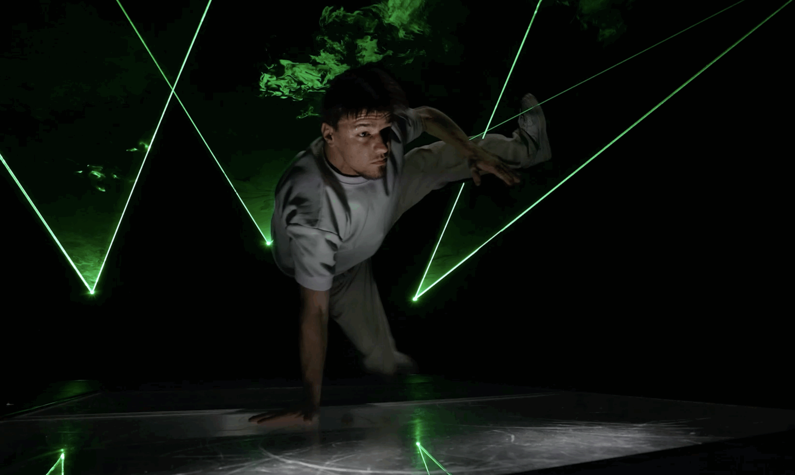 Ein Mann in einem grauen Outfit führt auf einer beleuchteten Bühne eine einhändige Breakdance-Bewegung vor, umgeben von grünen Laserstrahlen und dunklem Rauch im Hintergrund. Laser Dance Show