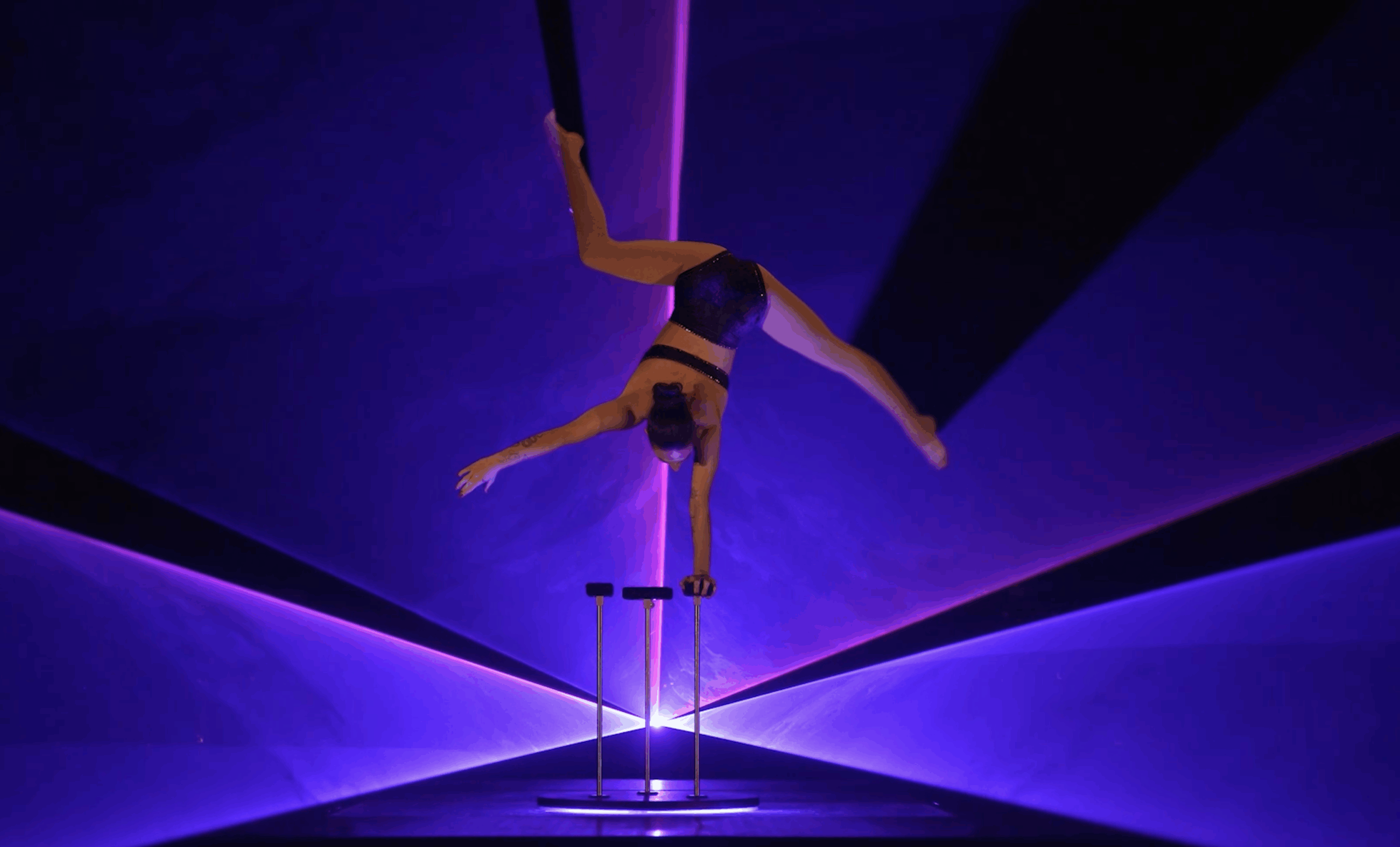 Laser Dance Show. Eine Person macht einen einarmigen Handstand auf Stöcken, die Beine in der Luft gespreizt, umgeben von dramatischer lila und blauer Bühnenbeleuchtung und Laserstrahlen.