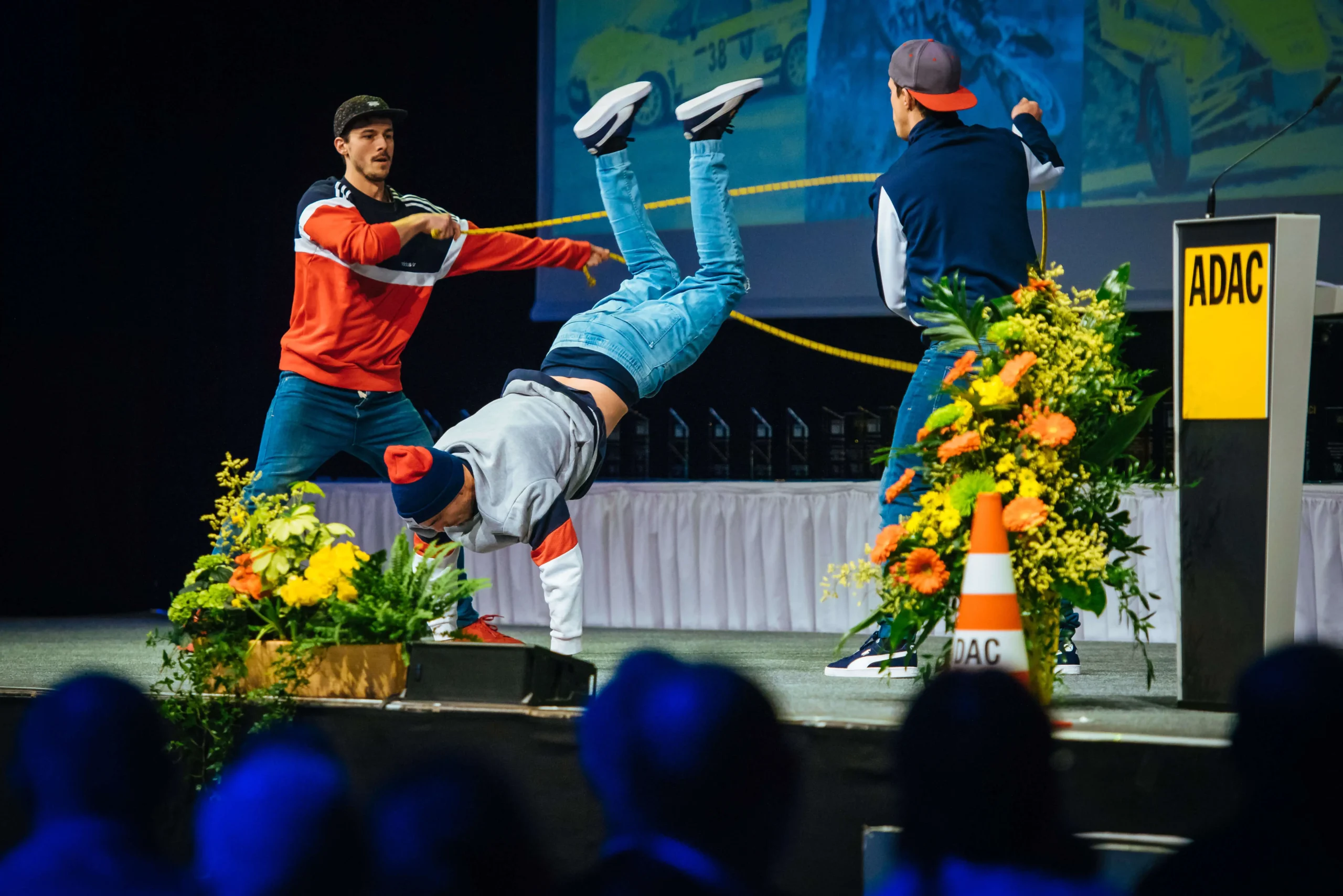 Drei Männer in legerer Straßenkleidung führen auf der Bühne eine tänzerische oder akrobatische Bewegung auf, die an eine Ropeskipping-Show erinnert; einer liegt kopfüber auf den Händen, während die anderen assistieren. Blumen und ADAC-Zeichen stehen im Vordergrund, während das Publikum zusieht.