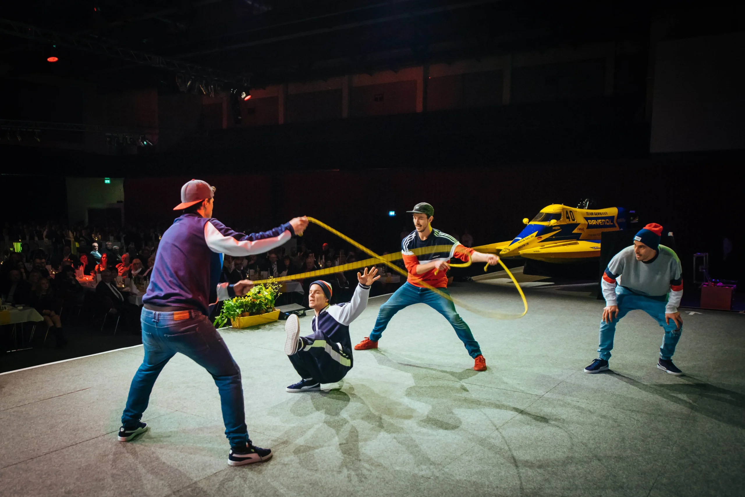 Vier Personen führen auf der Bühne eine Ropeskipping-Show auf; zwei halten ein langes gelbes Seil, während eine dritte Person in der Mitte hockt und springt. Die vierte Person sieht zu, wie das Publikum und ein großes gelbes Boot im Hintergrund zu sehen sind.