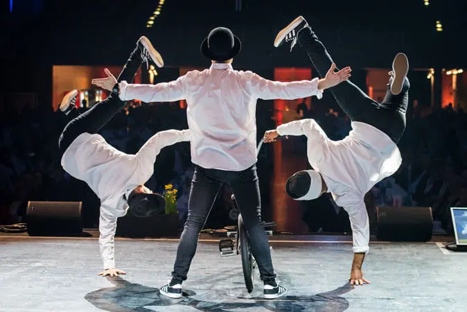 Drei Tänzerinnen und Tänzer treten auf der Bühne in einer Mix-Show auf; die mittlere Tänzerin oder der mittlere Tänzer steht mit ausgestreckten Armen dem Publikum zugewandt, während die anderen auf beiden Seiten einhändige Handstände machen, alle gekleidet in weiße Hemden, schwarze Hosen, Hüte und Turnschuhe.