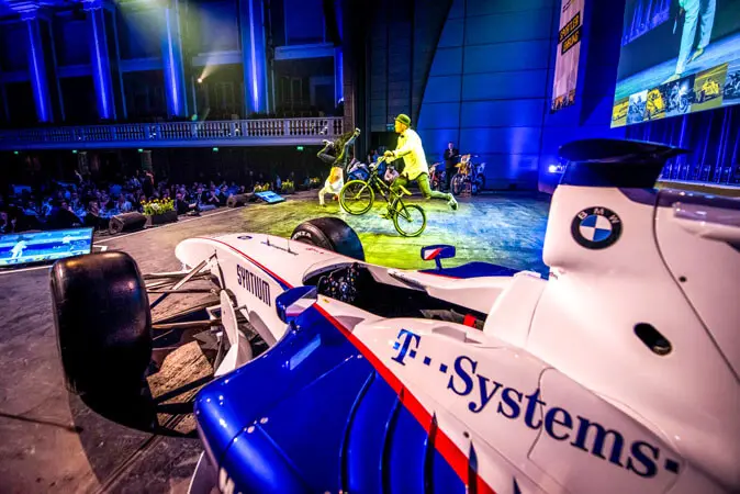 Ein weiß-blaues BMW Formel-1-Auto wird während einer Mix-Show in einem Innenraum auf einer Bühne ausgestellt, während zwei Radfahrer in der Nähe Kunststücke vorführen. Der Raum ist mit blauen Scheinwerfern hell erleuchtet und ein Publikum schaut aus dem Hintergrund zu.
