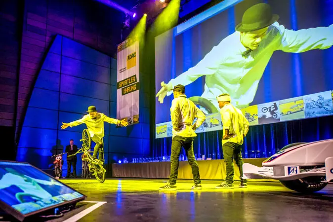 Ein Künstler in weißem Hemd und Melone balanciert während einer Mix-Show auf einem Fahrrad, während zwei andere zuschauen. Ein großer Bildschirm im Hintergrund zeigt eine Nahaufnahme der Darbietung mit stimmungsvoller Beleuchtung und dem im Hintergrund sichtbaren Publikum.